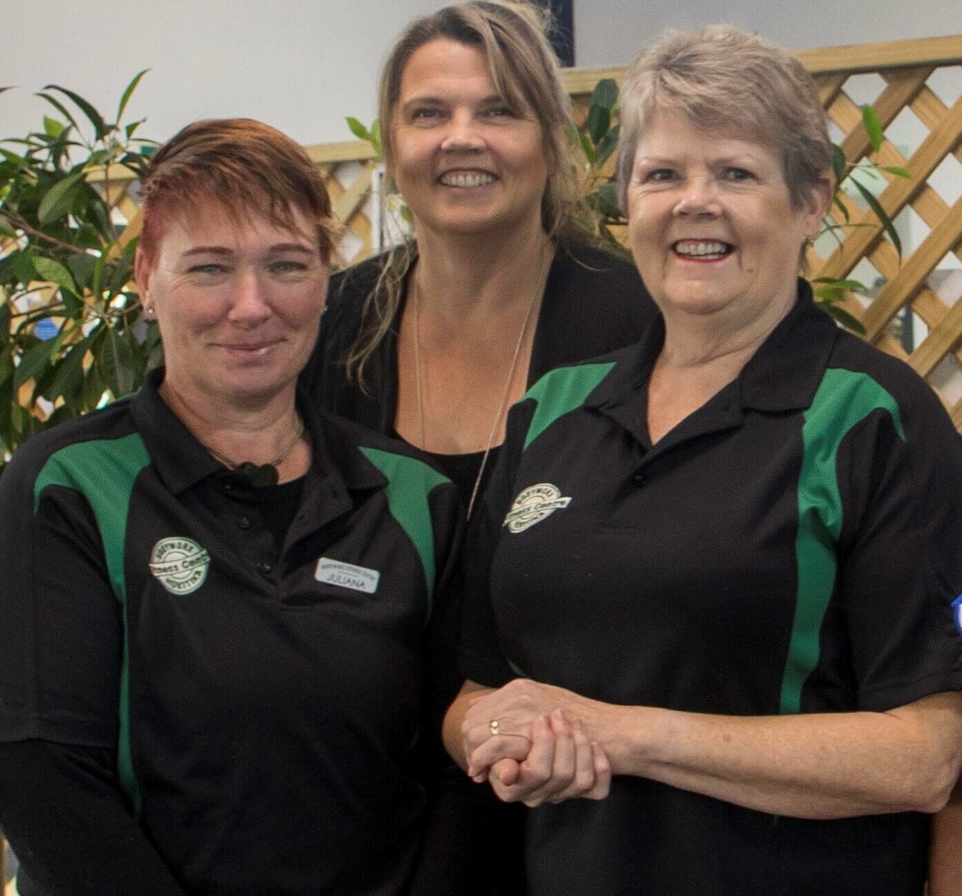Three women standing together, two wearing matching black and green collared shirts with a logo, the middle woman in a black top. They are smiling and posing for the photo in front of a plant.