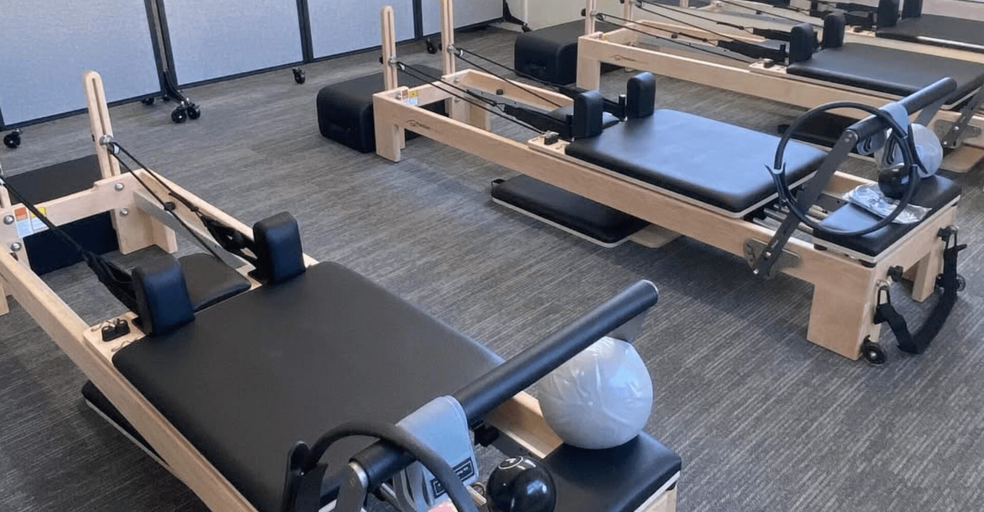 10 Several Pilates reformer machines with black padding and wooden frames are arranged in a fitness studio with grey carpeted flooring.