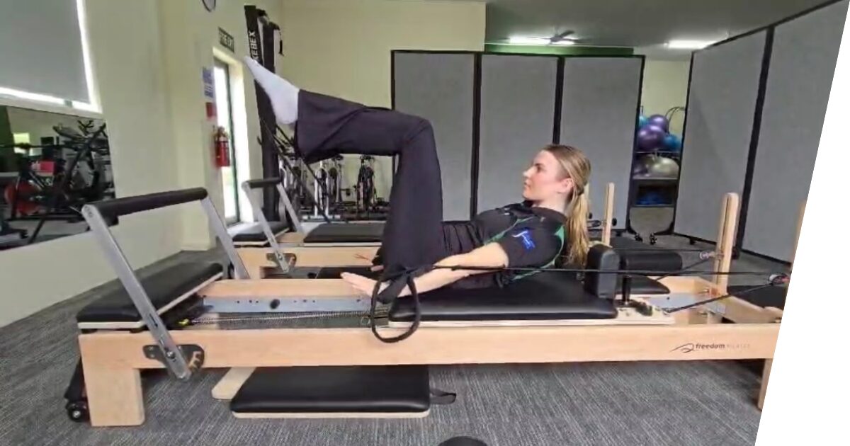 A woman performs a Pilates exercise on a reformer machine in a gym, lying on her back with her legs raised and bent at a right angle.